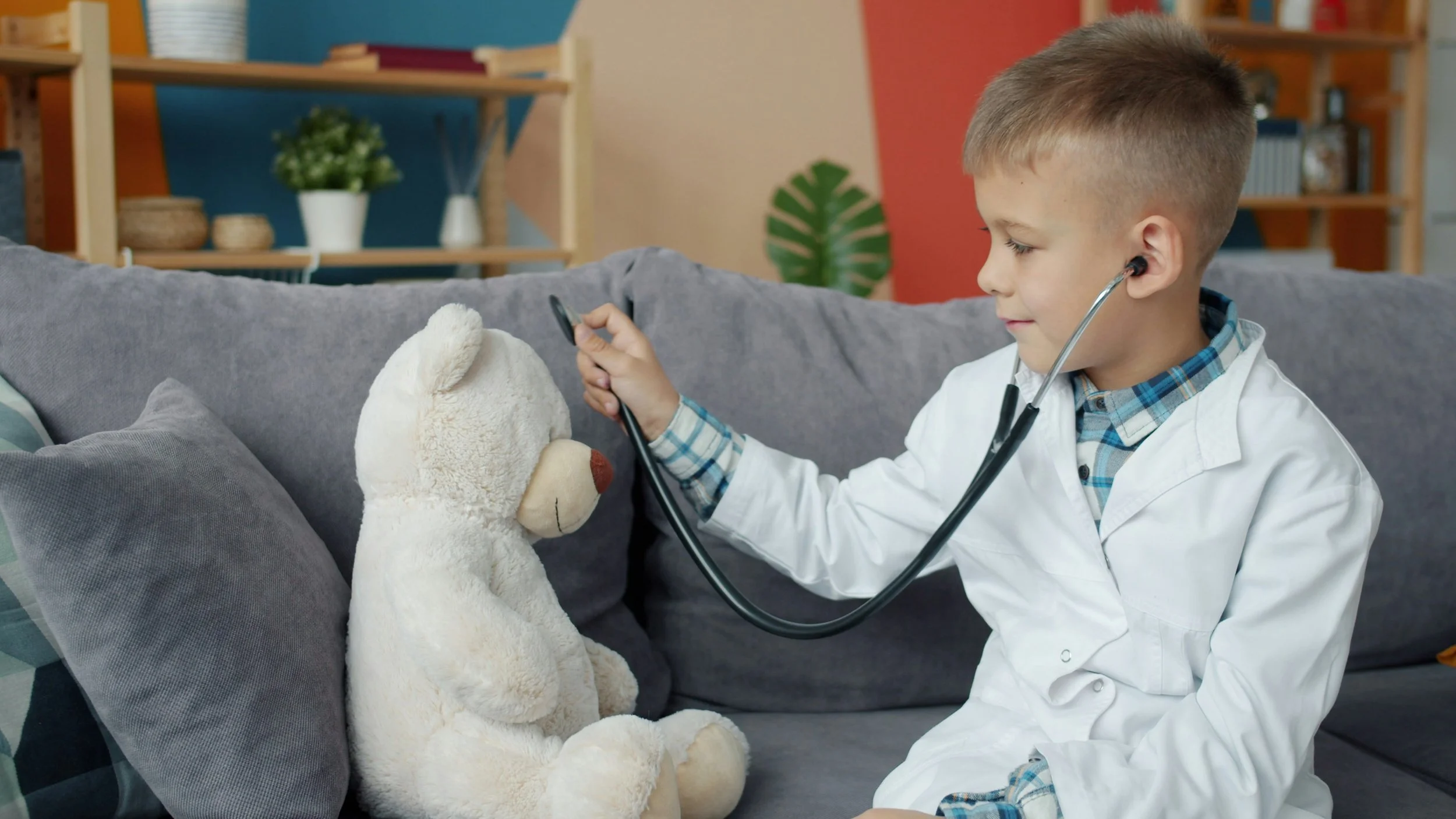Child dressed as a doctor using a stethoscope to listen to a teddy bear's heartbeat on a couch.