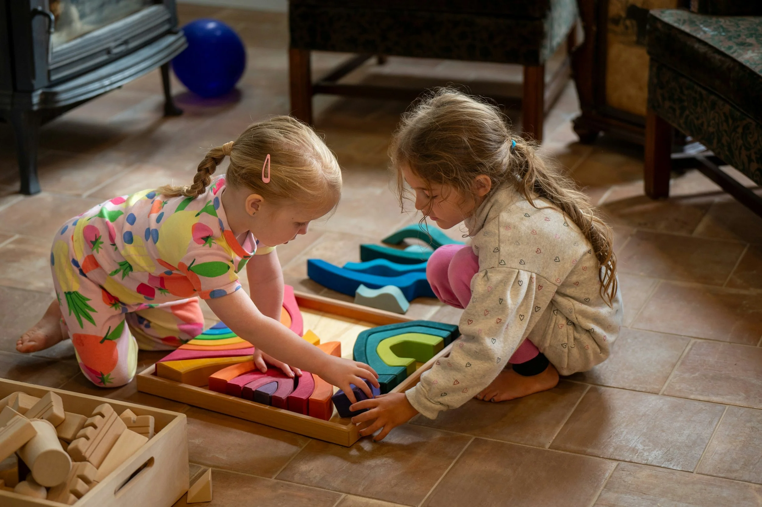 Two young girls playing with wooden rainbow stacking blocks on the floor, with furniture and a blue ball in the background.