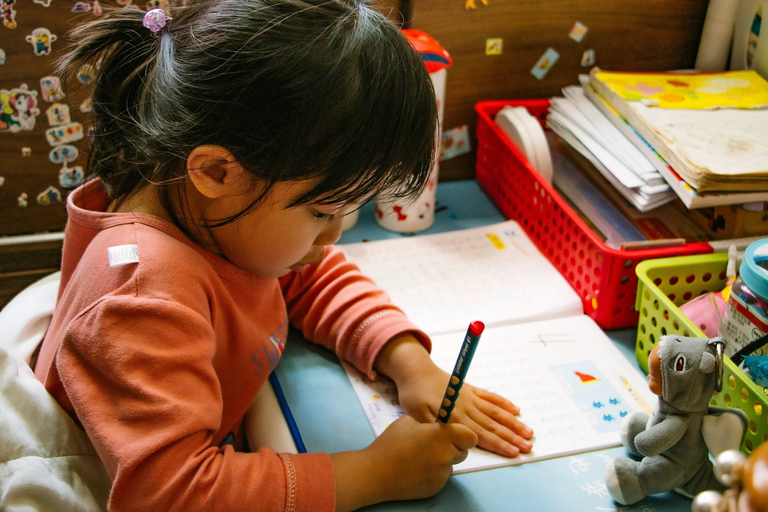 A young girl with black hair, wearing a pink sweatshirt, sitting at a desk writing in a notebook with a pencil. The background features a wooden wall decorated with colorful stickers, a red basket filled with books and papers, and a yellow folder. There are also various toys and stationery items on the desk.
