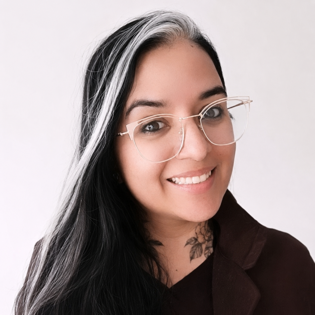 A woman with long black hair with silver streaks, wearing large clear glasses, smiling, and a tattoo of a flower on her neck, against a plain white background.