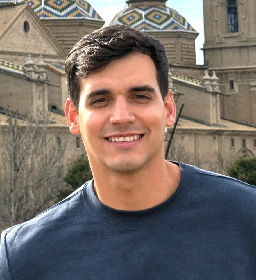 A young man with dark hair smiling outdoors in front of a historic building with colorful tiled domes.