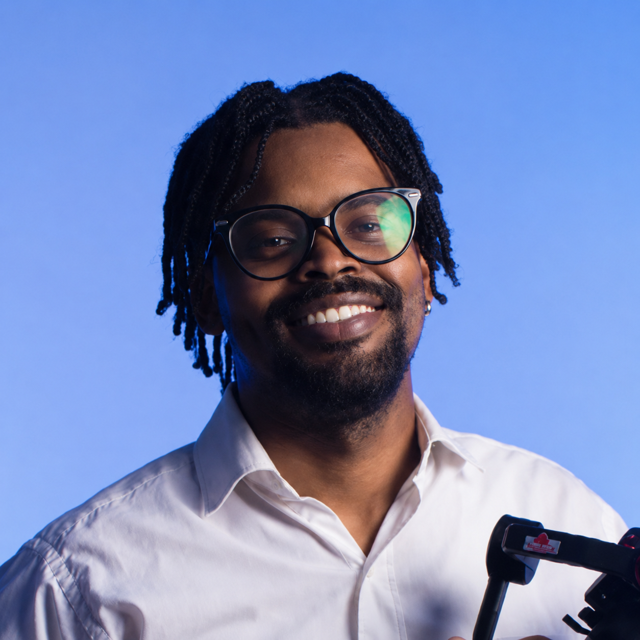 A smiling man with glasses and dreadlocks wearing a white shirt against a blue background.
