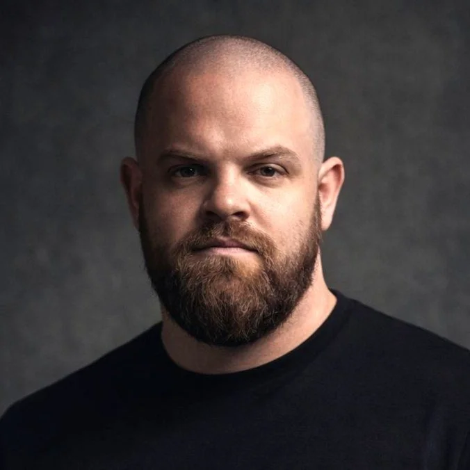 A portrait of a bald man with a beard, wearing a black shirt, against a dark background.