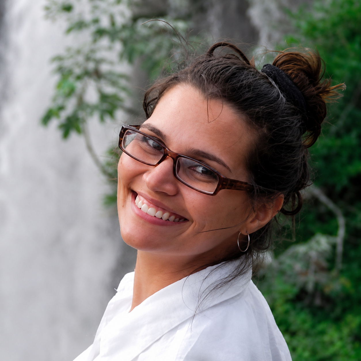 A woman with glasses and hoop earrings smiling outdoors near a waterfall with greenery.