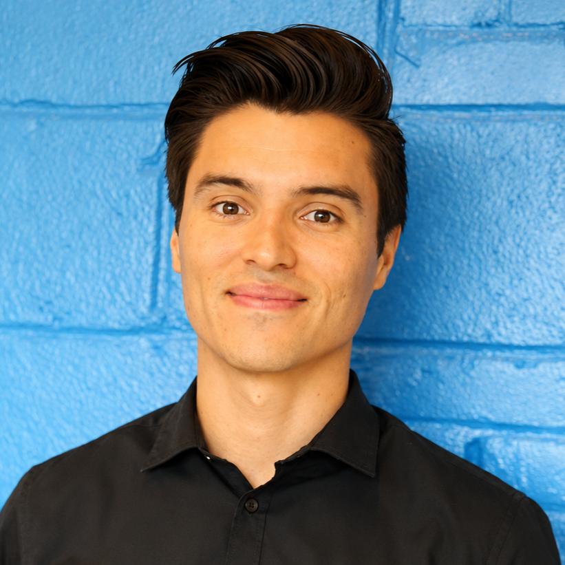 Portrait of a young man with dark hair and fair skin, wearing a black collared shirt, standing against a bright blue textured wall.