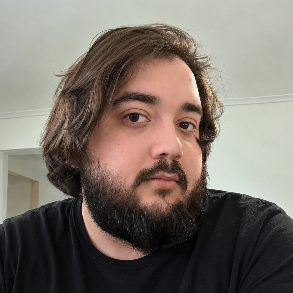 Close-up photo of a person with shoulder-length brown hair and a full beard, wearing a black shirt, in a room with light-colored walls.