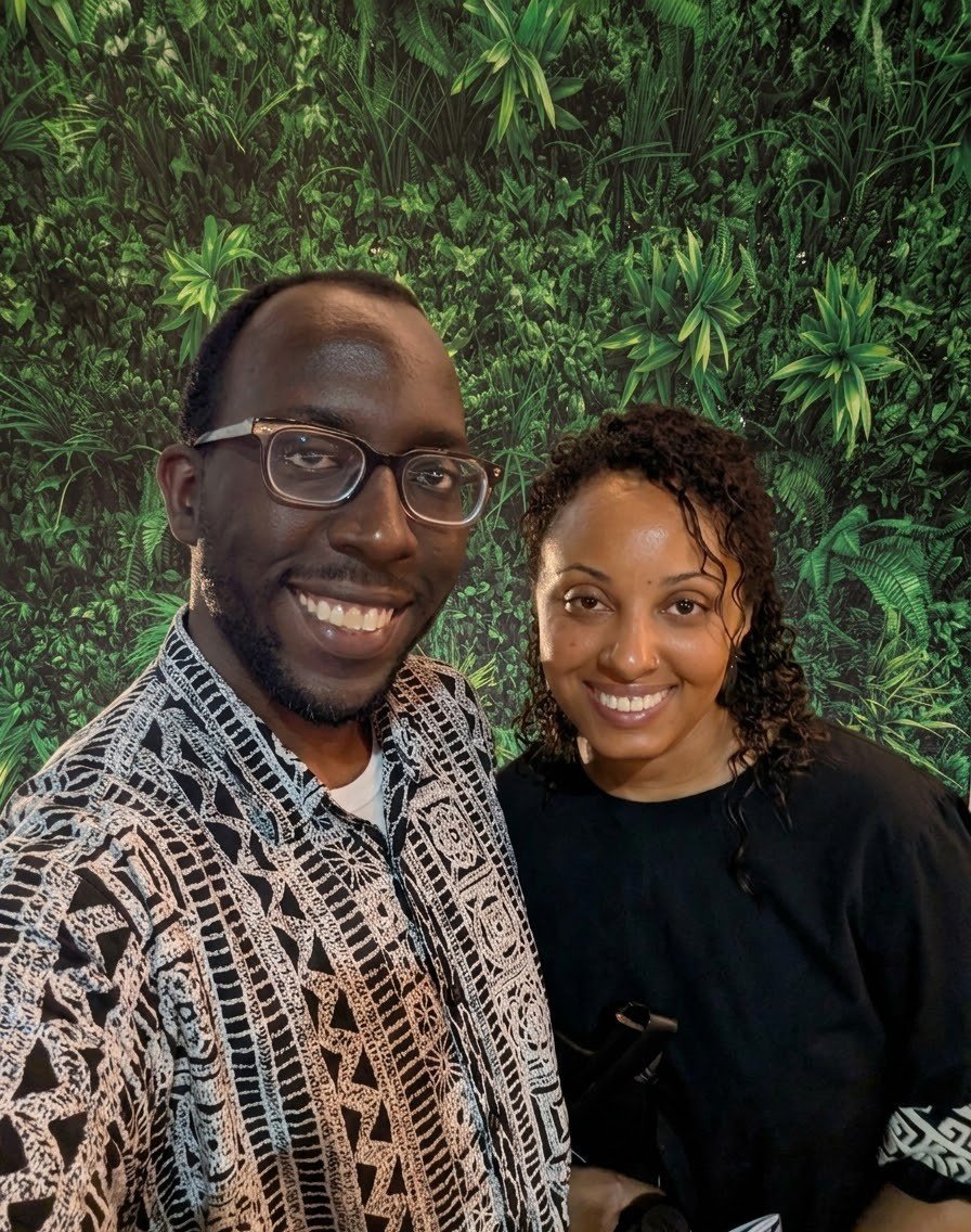 Two smiling people, a man with glasses and a woman, standing in front of a green leafy jungle-themed backdrop.