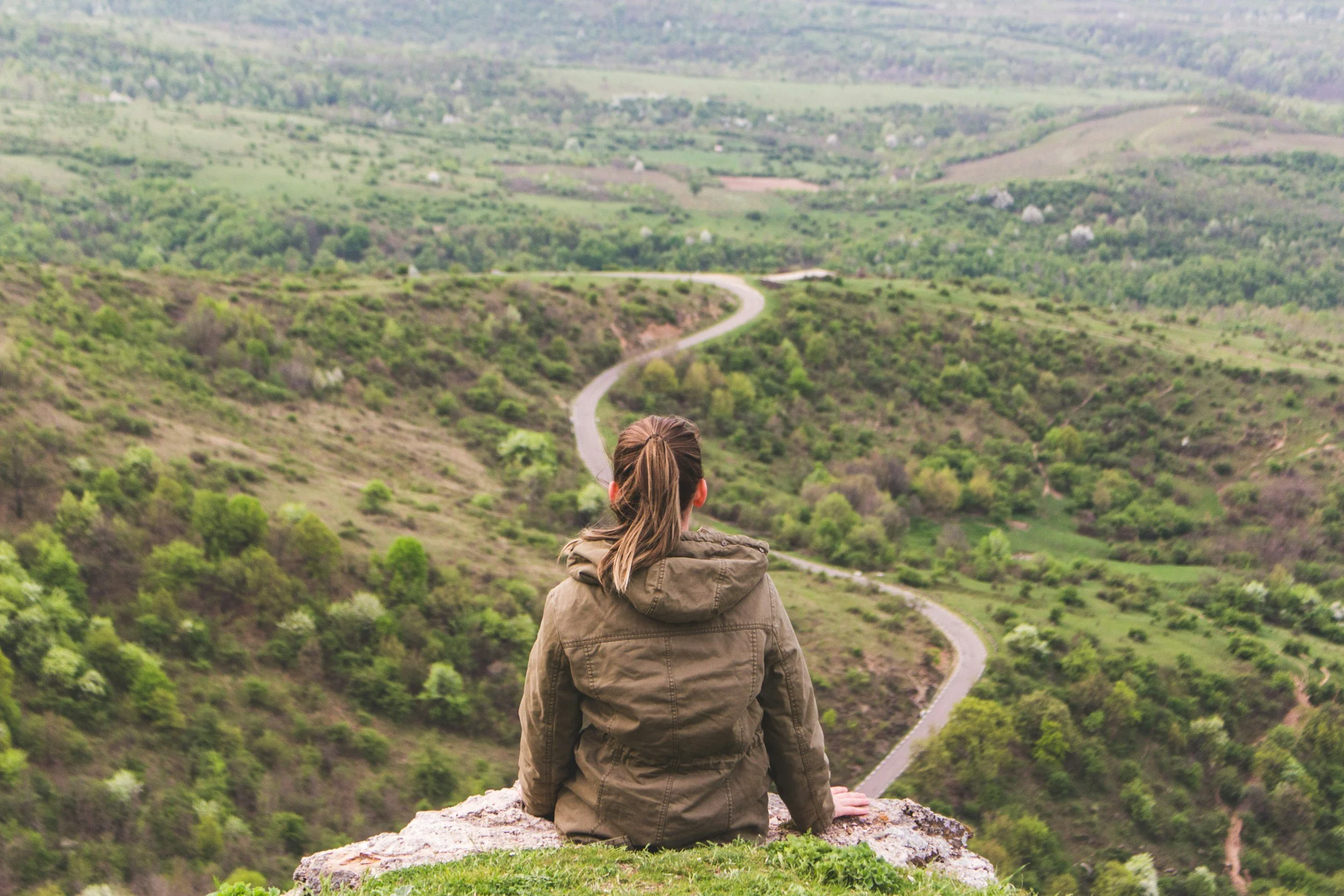 A person with brown hair in a ponytail wearing a brown jacket, sitting on a rock, overlooking a winding road through green hills and sparse trees.