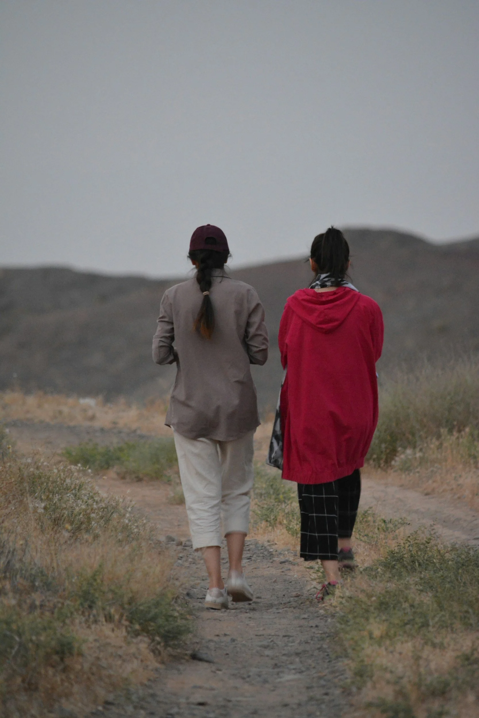 Two women walking on a dirt trail in a desert landscape, dressed casually, with mountains in the background during dusk or dawn.