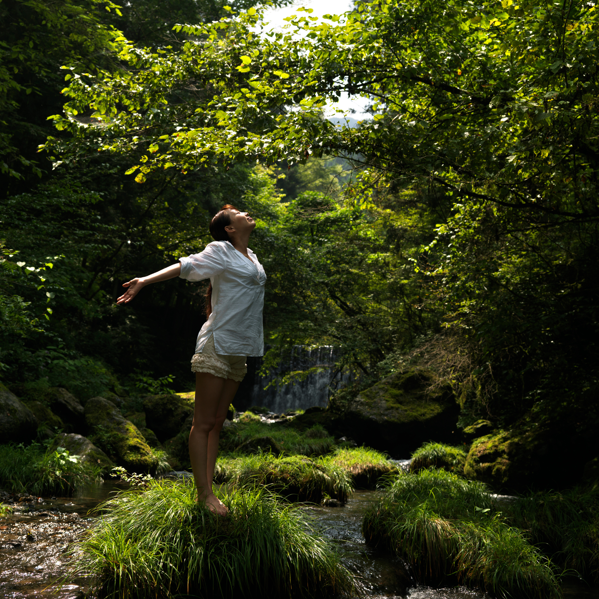 A woman in a white shirt and beige shorts stands with arms outstretched in a lush, green forest during daytime, near a small stream with a waterfall in the background, enjoying nature.