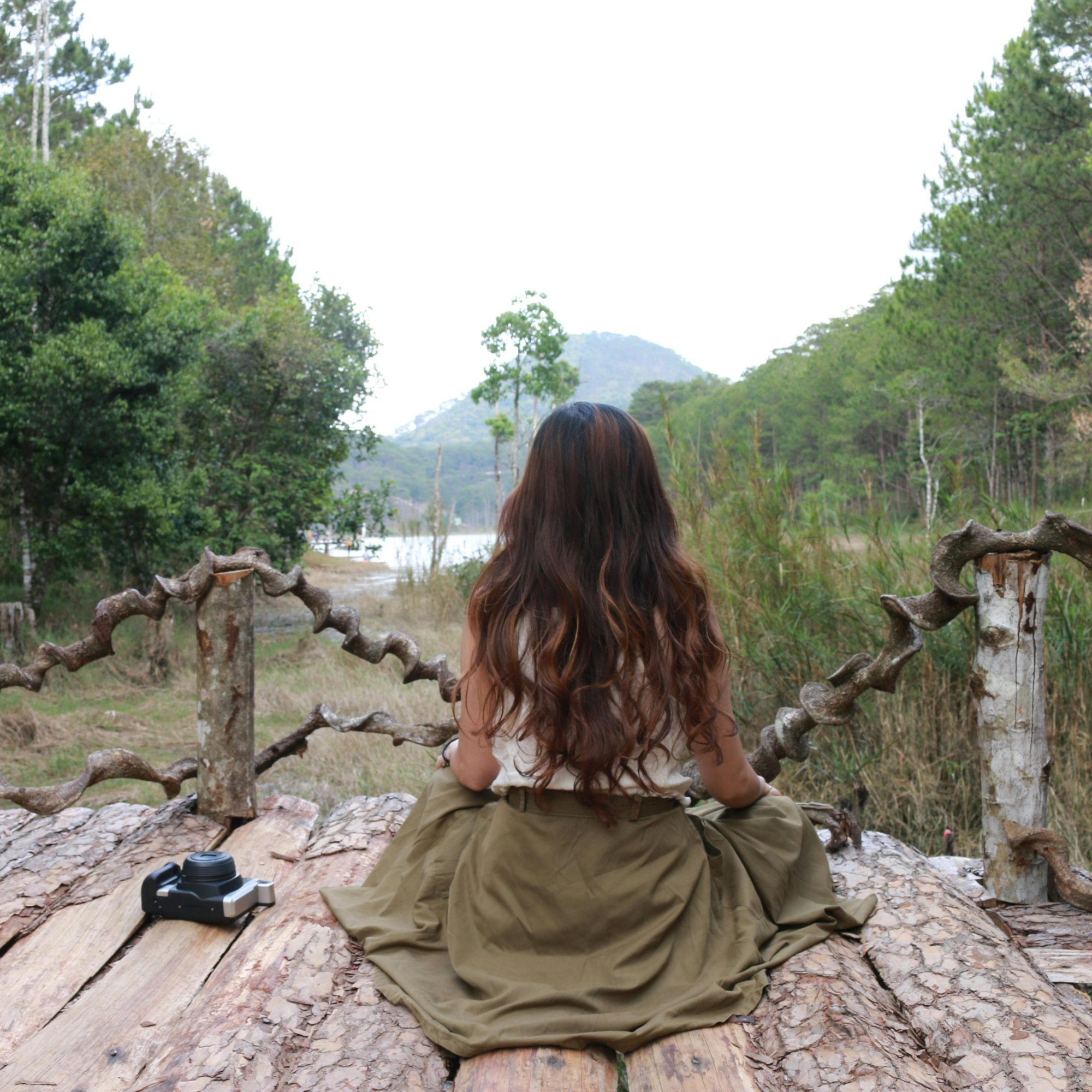 A woman with long brown hair sitting cross-legged on a wooden platform outdoors, facing away, overlooking a forested landscape with trees and mountains overcast sky in the background.