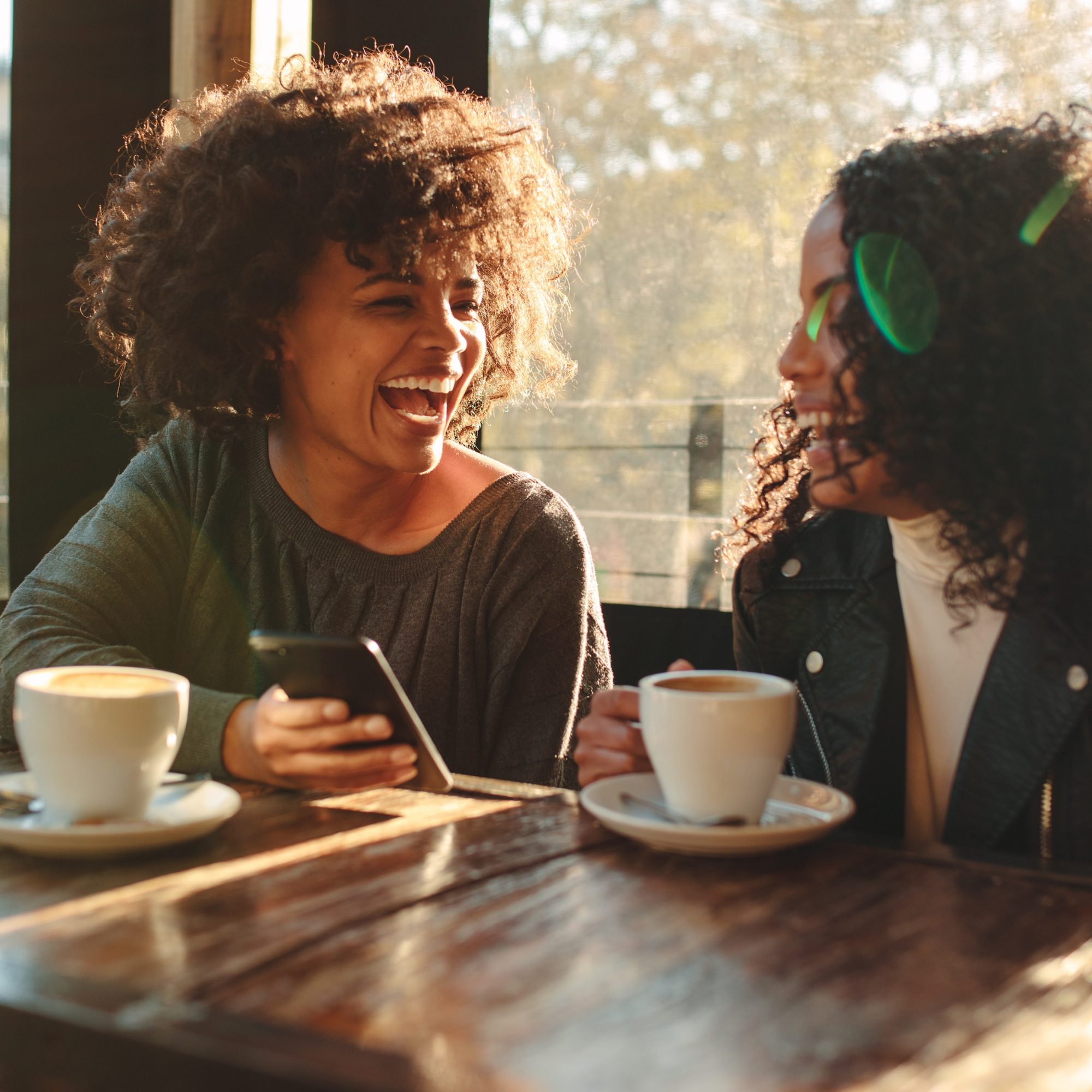 Two women are sitting in a cafe, smiling and enjoying each other's company. One woman is holding a smartphone, and both have coffee mugs on the table in front of them. Sunlight is coming through the window behind them.
