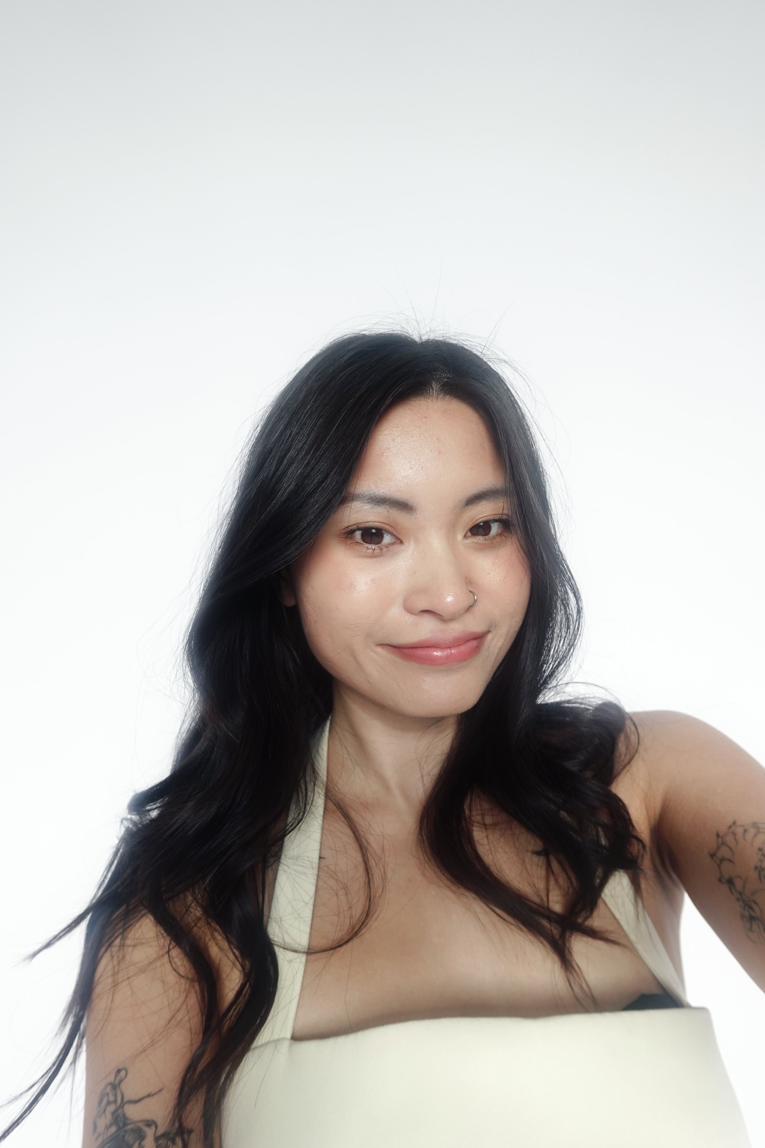 A woman with black wavy hair, wearing a beige tank top, taking a selfie against a plain white background.