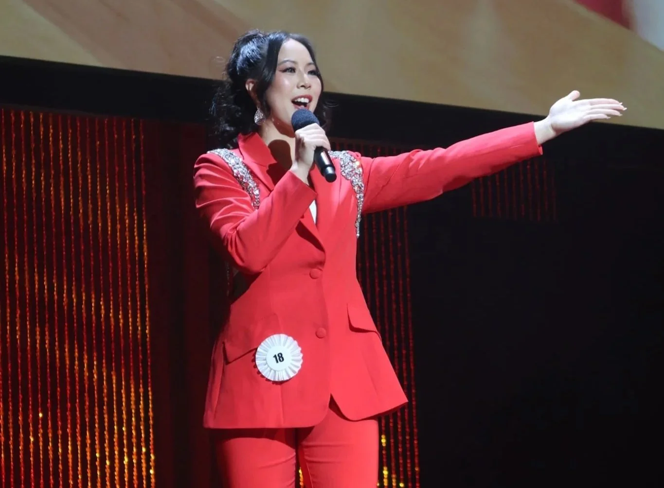 A woman in a red suit with embellished shoulders and a badge number 18 is speaking into a microphone on a stage with a dark background.