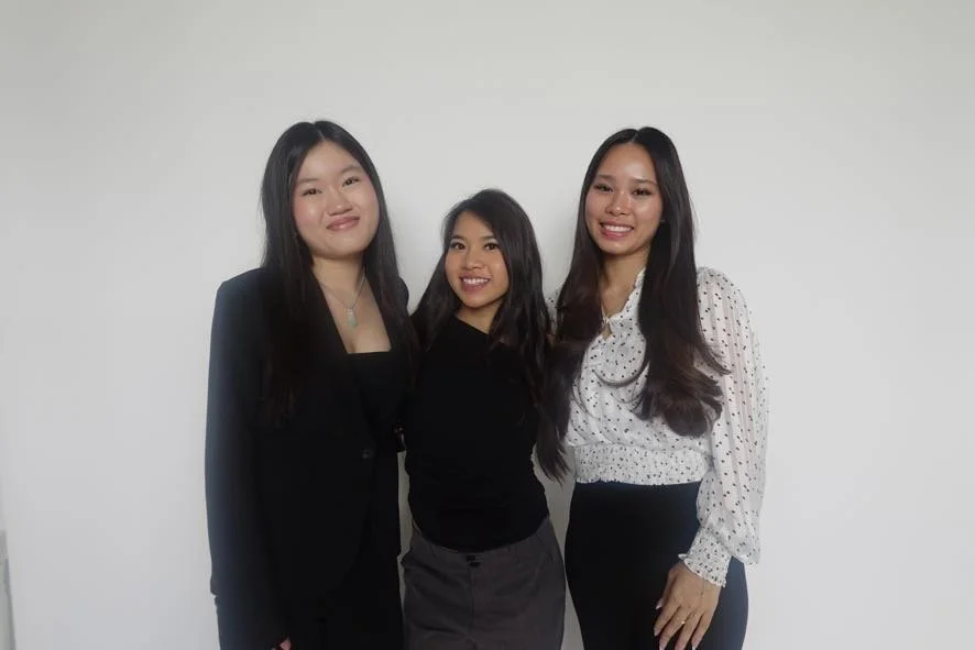 Three young women standing together, smiling against a plain white wall. They are dressed professionally, with long dark hair.