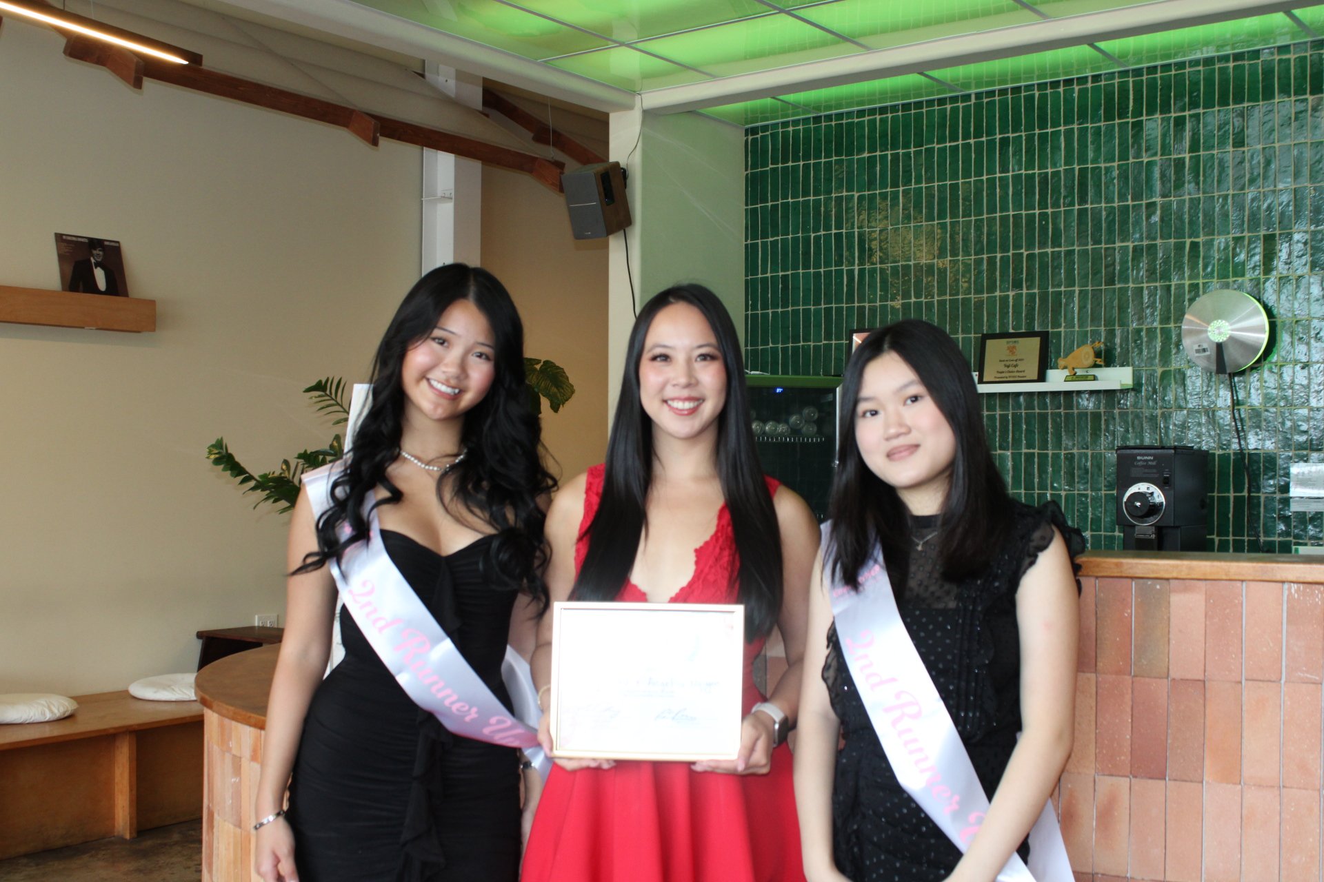 Three women standing together indoors, two wearing sashes that say 'Miss Girlz Queen' and one holding a certificate, all smiling for the photo.