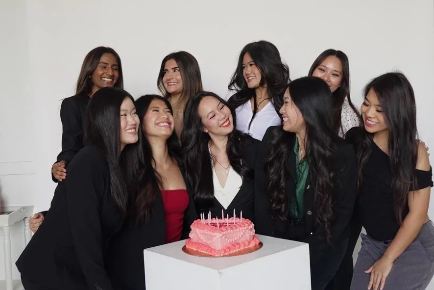 A group of nine women celebrating a birthday with a pink cake in front of them.