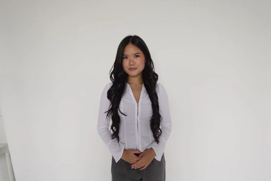 A young woman with long black hair, wearing a white shirt and gray pants, standing against a plain white background.