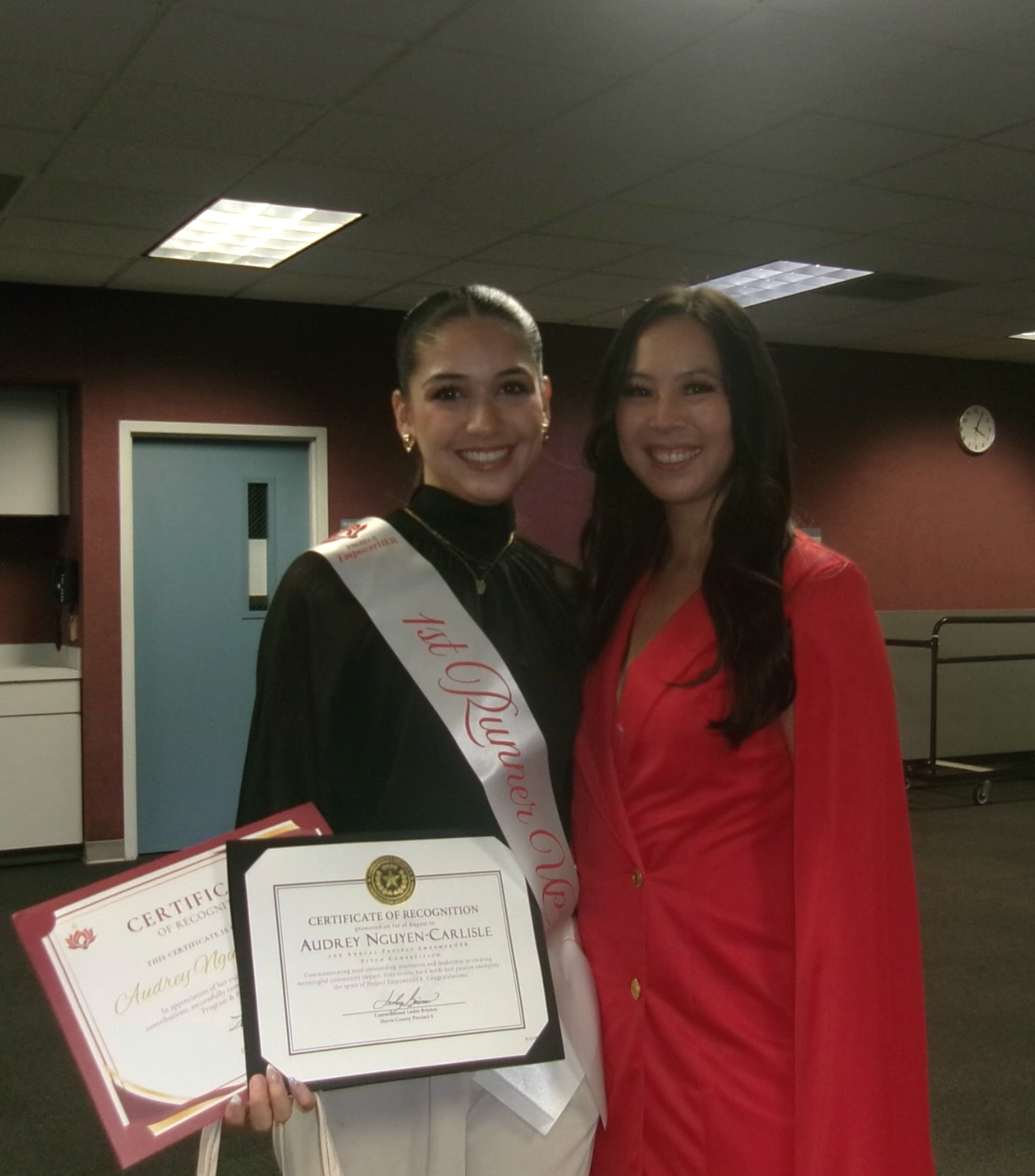 Two women smiling, one wearing a graduation gown with a sash and holding certificates, the other in a red dress, standing indoors in a room with ceiling lights and a clock.