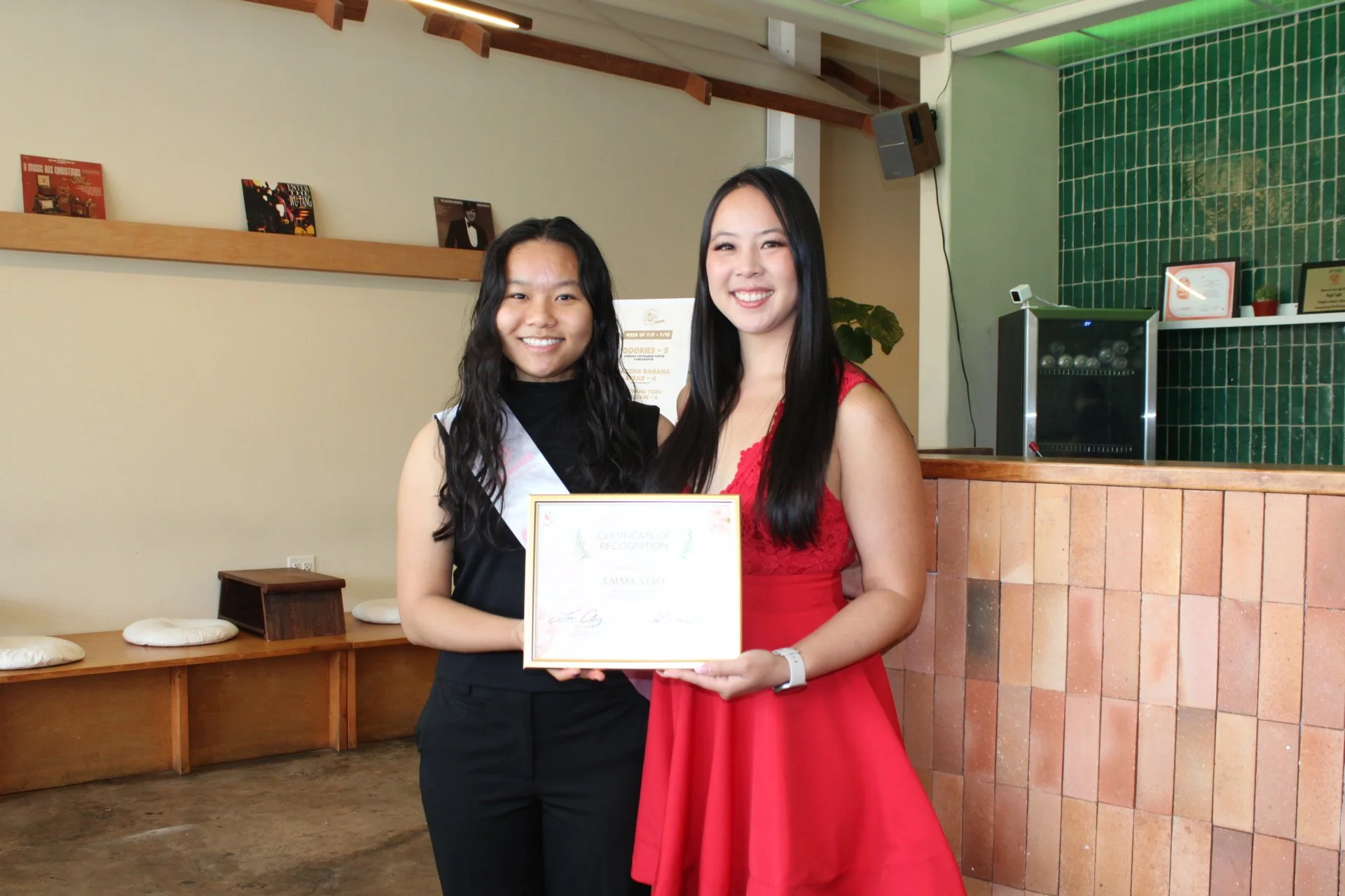 Two women standing inside a cafe or restaurant, smiling and holding a certificate of recognition. One woman wears a black dress with a sash, and the other wears a red dress. Behind them, there are green tiled walls and a counter with framed certificates and a small refrigerator.
