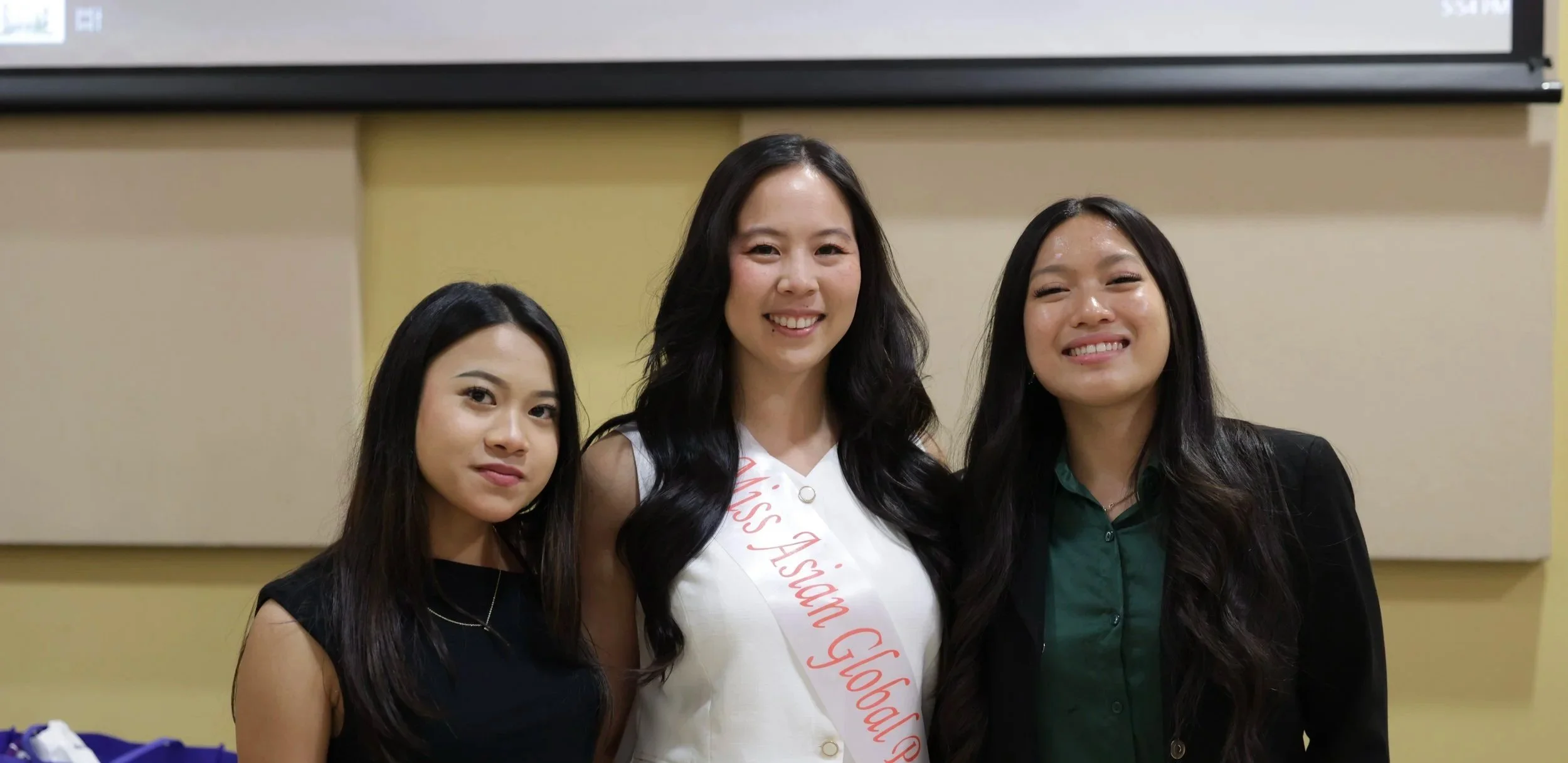 Three young women smiling, standing side by side indoors. The woman in the middle is wearing a sash that says 'Miss Asian GLOBA' and has long black hair. The woman on the left also has long dark hair and wears a black top, while the woman on the right has long dark hair, wearing a dark blazer and green shirt.
