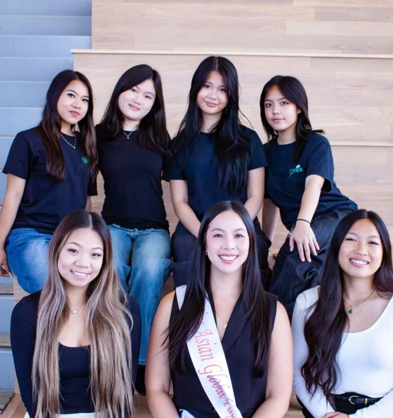 A group of young women posing together for a photo in a casual setting with wooden wall background. One woman in the front is wearing a sash that says "Asian G” and is smiling. Others are standing and sitting behind her, all smiling and dressed casually.