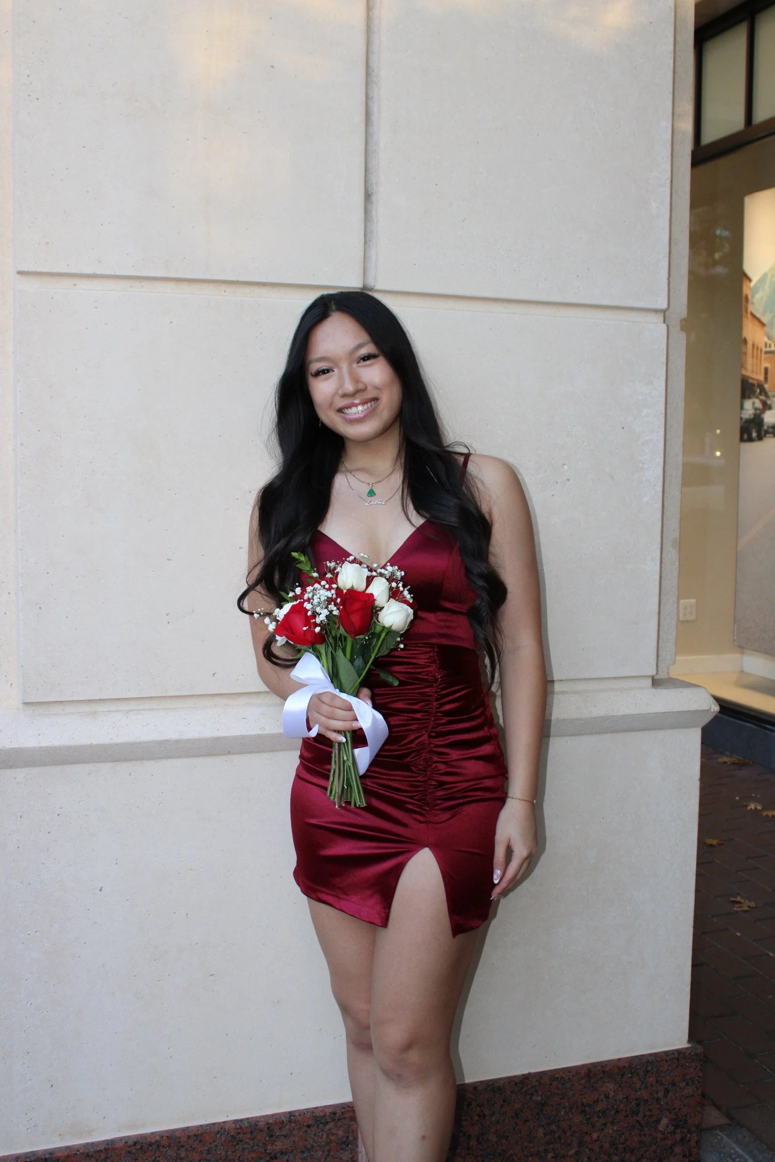 Young woman in a burgundy dress holding a bouquet of red and white roses, standing against beige wall with glass window to the right.