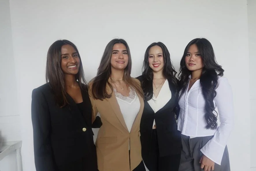 Four women standing together indoors, smiling, dressed in professional attire.