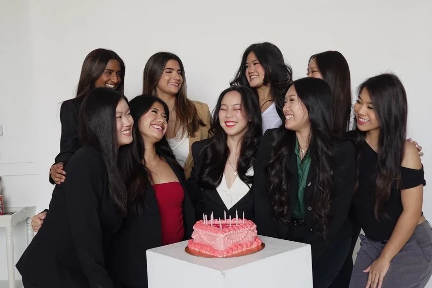 Group of women celebrating around a pink birthday cake with candles.