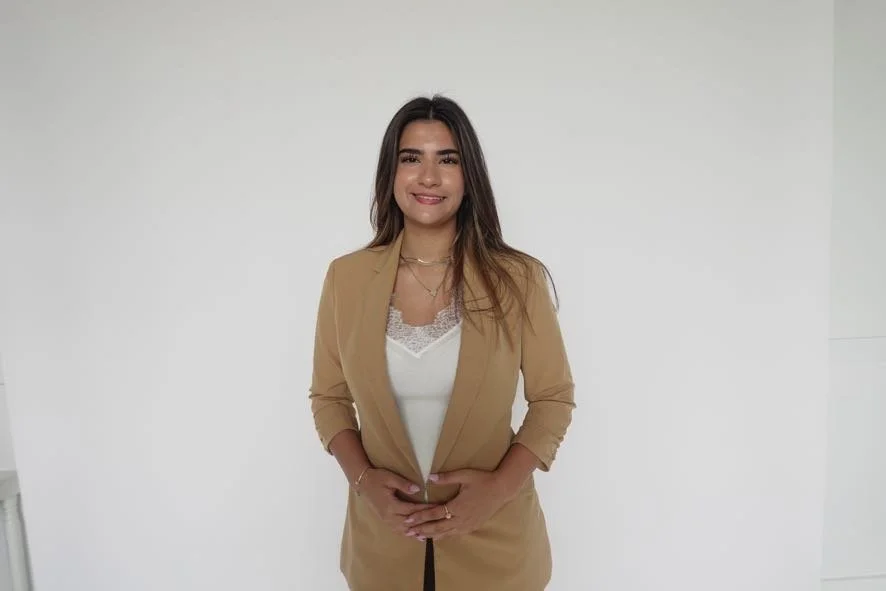 A young woman with long brown hair, smiling, standing against a white background, wearing a beige blazer over a white top.