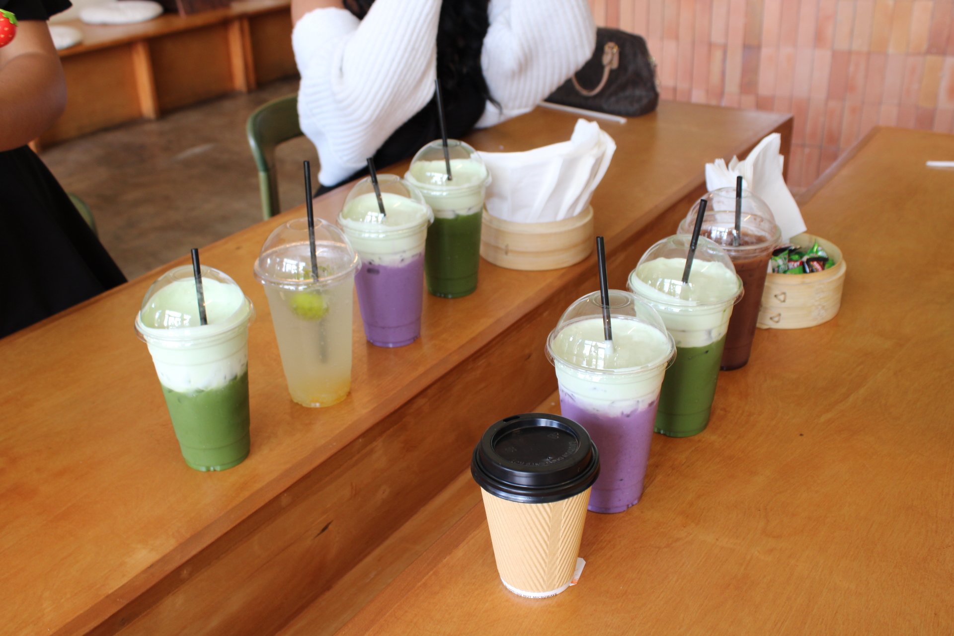 Assorted colorful smoothies and drinks on a wooden table, with some in clear cups with dome lids and straws, and one in a paper coffee cup. A basket of small candies or snacks and paper napkins are also visible.