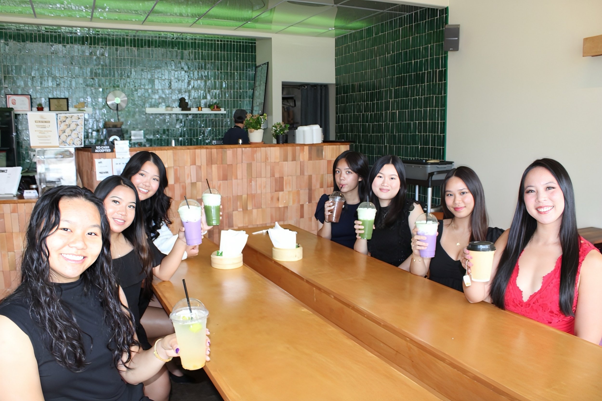 A group of friends sitting at a wooden table in a cafe, holding colorful drinks and smiling at the camera, with a green tiled wall in the background.
