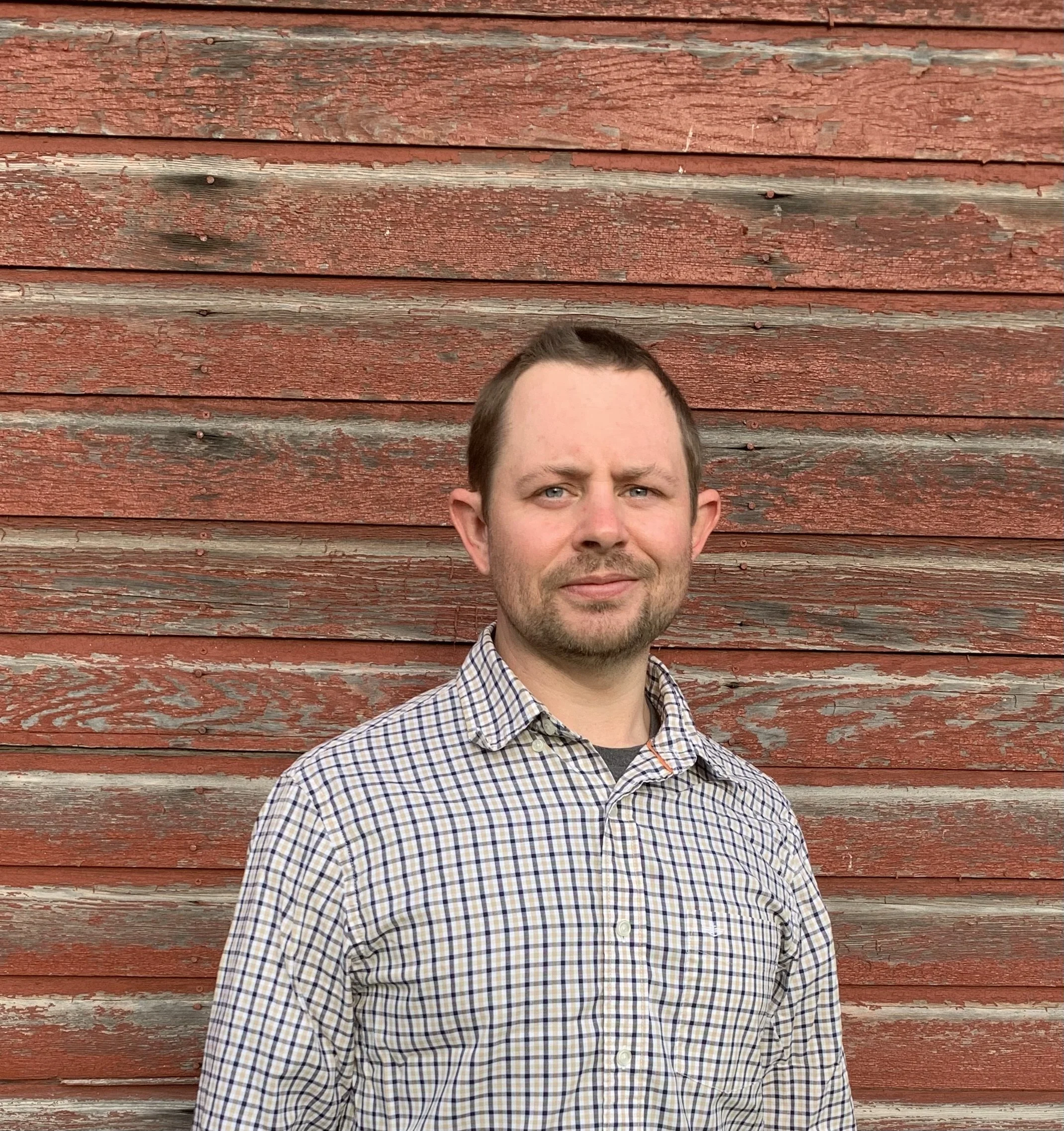 A man with short brown hair and a light beard wearing a checkered shirt standing in front of a weathered red wooden wall.