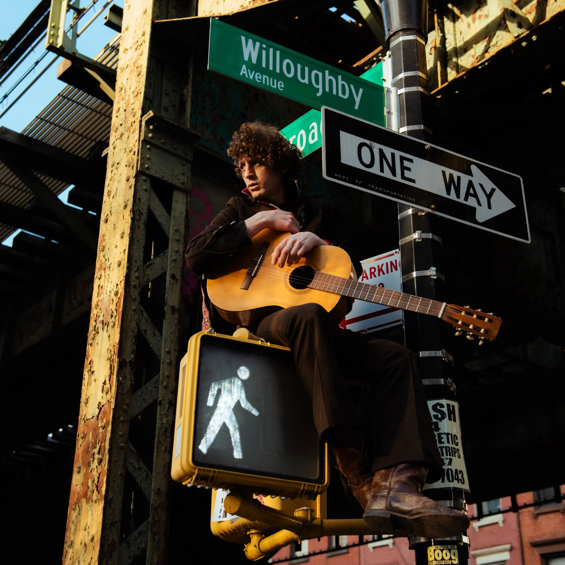 Adam Amram playing an acoustic guitar on a street corner under street signs for Willoughby Avenue and another street, with a pedestrian crosswalk signal below them.