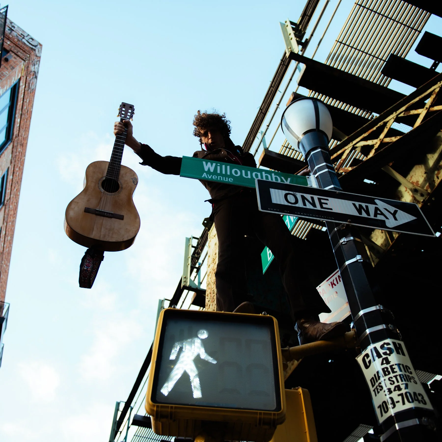 Street scene with a person holding a guitar, a pedestrian crossing signal showing a walking figure, street signs reading 'Willoughby Avenue' and 'One Way', and a partially visible pole with advertising stickers, taken from a low angle during daytime.