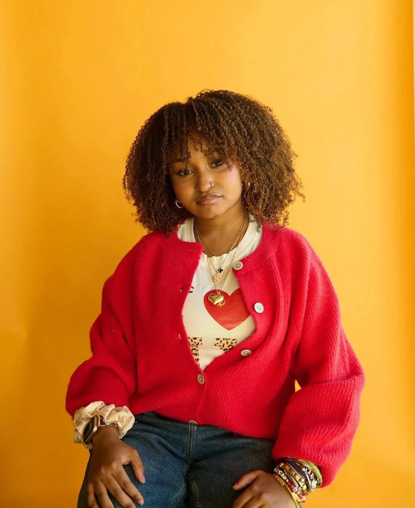 A young woman with curly hair wearing a red cardigan with large buttons, layered jewelry, and a graphic t-shirt, sitting against a yellow background.