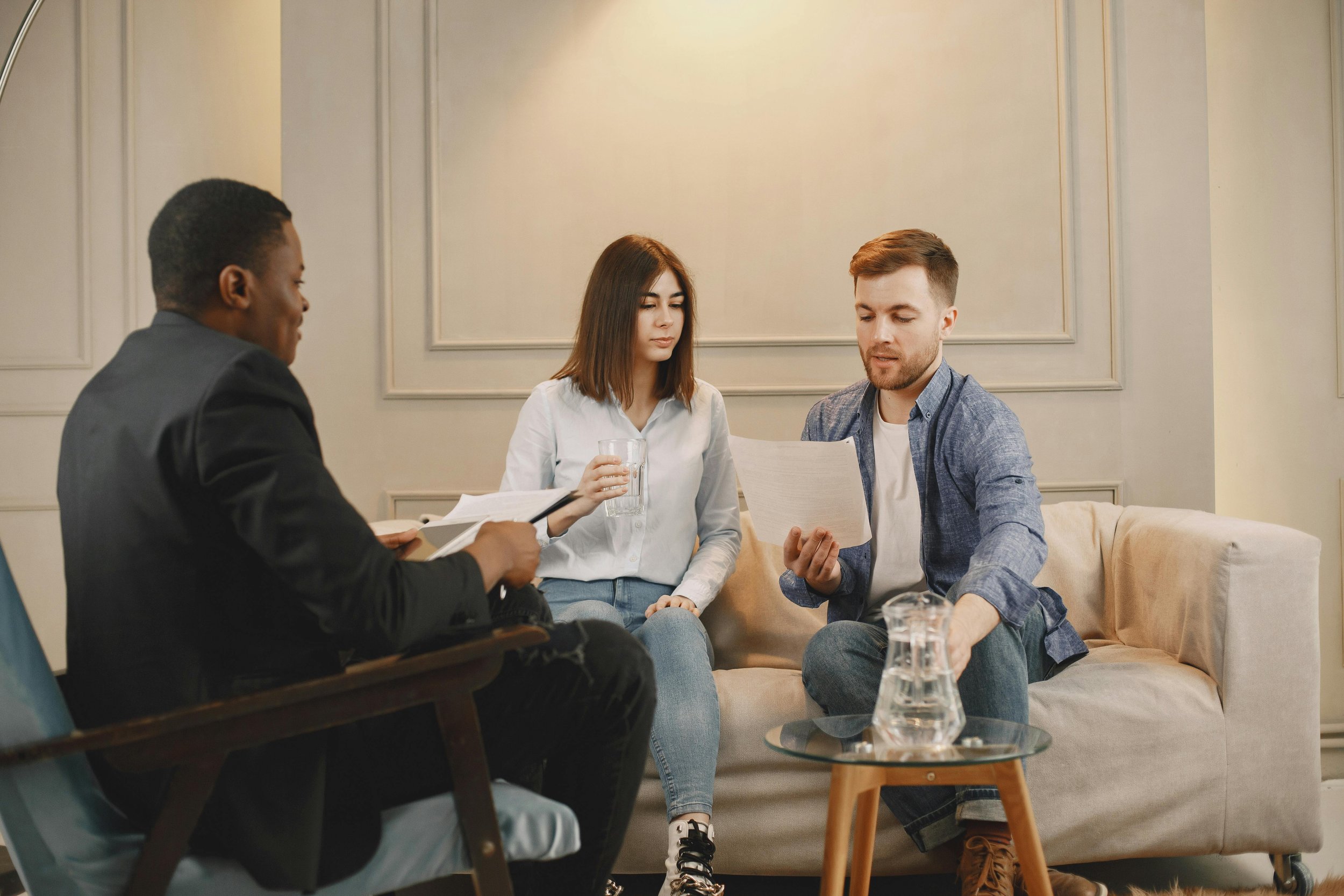 A young man and woman sitting on a couch, engaged in a discussion with a middle-aged man, holding papers and a glass of water.
