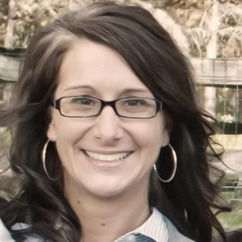 A woman with dark hair, glasses, and hoop earrings smiling outdoors with trees and a fence in the background.