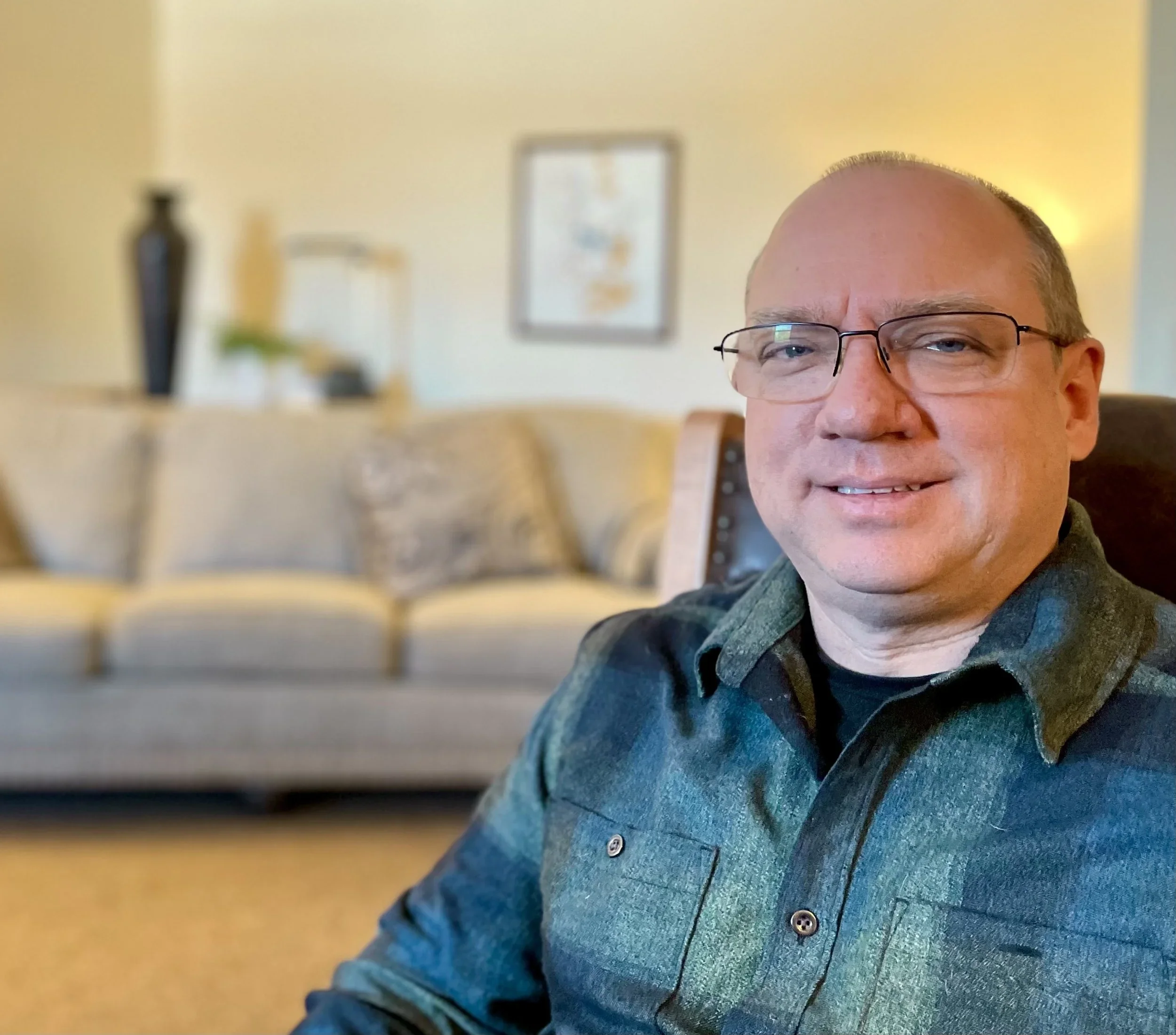 A smiling man wearing glasses and a dark-colored shirt with a textured pattern, sitting indoors with a blurred yellow wall, a framed picture, and a decorative vase in the background.