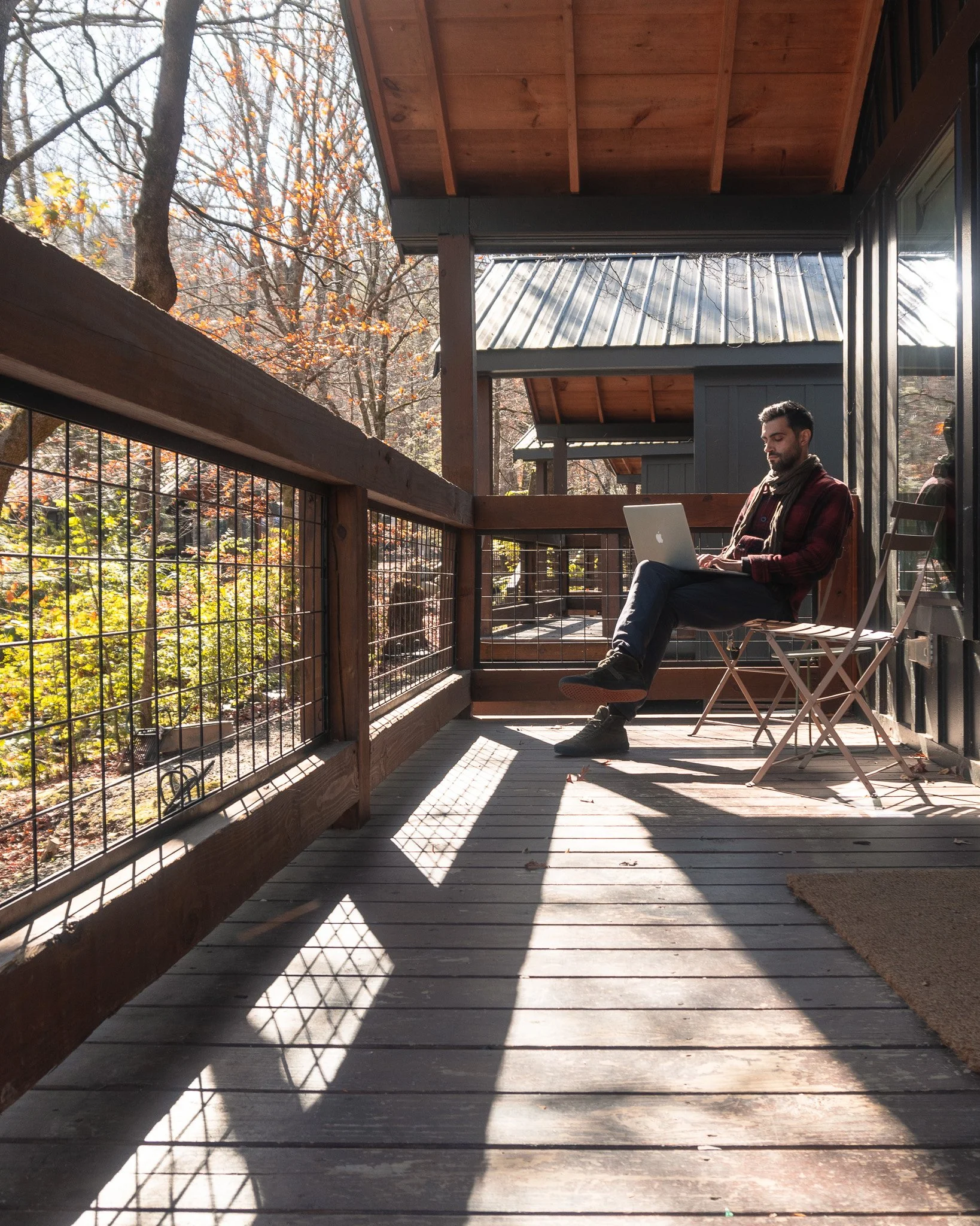 A man sitting on a wooden chair on a porch, working on a laptop, with trees and autumn foliage in the background.