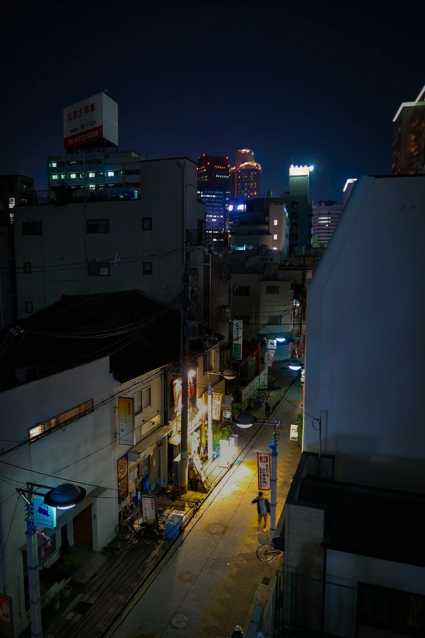 Nighttime city street view with buildings, neon lights, and a few pedestrians walking along the sidewalk.