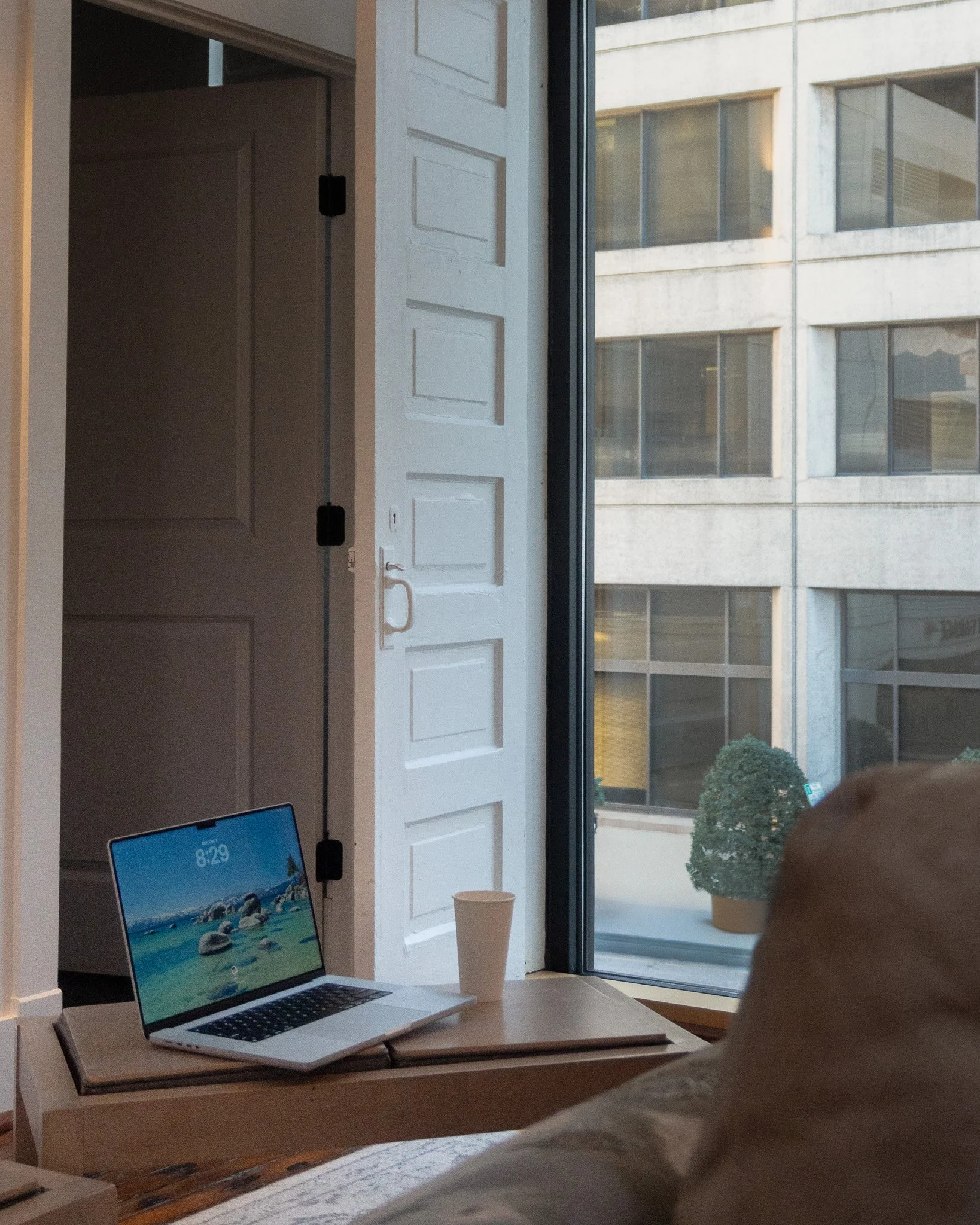 A computer on a desk with a quote paper cup, in front of a window overlooking a building with multiple windows.