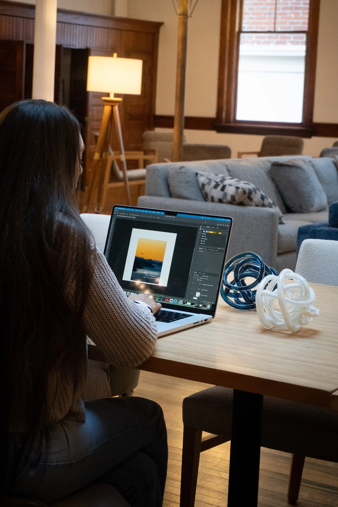 A woman with long dark hair sitting at a wooden table working on a laptop with nature-themed digital artwork on the screen. She is in a cozy living room with a sofa, decorative pillows, floor lamp, and a window with a brick building outside.