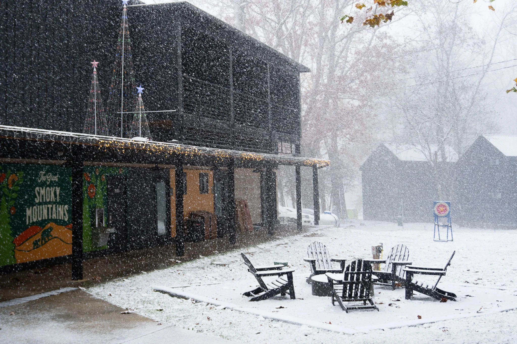 Snow falling on an outdoor patio area with black metal chairs and tables, a building decorated with Christmas lights and star decorations, and trees in the background with some leaves.