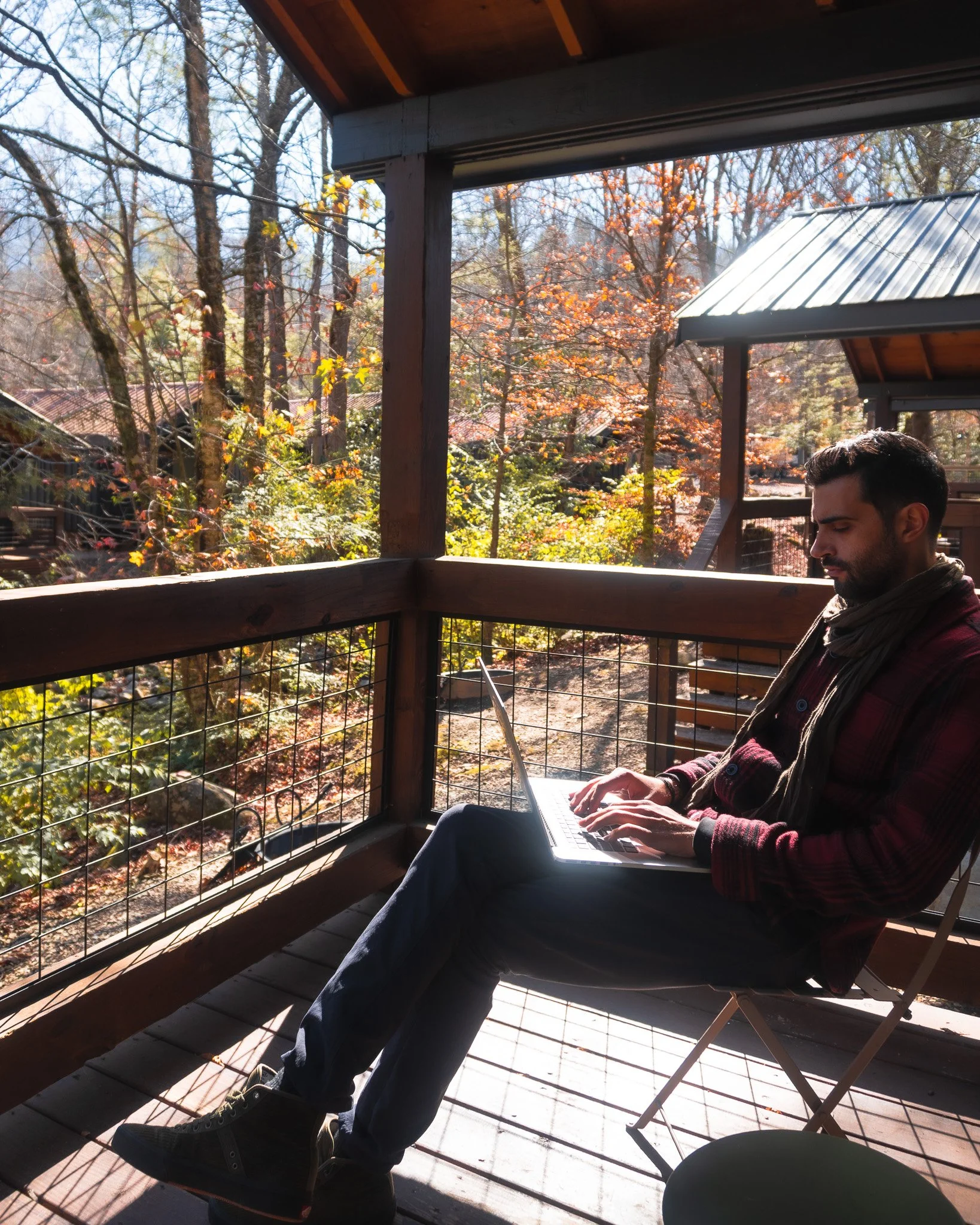 A man sitting on a deck with a laptop, surrounded by trees with autumn leaves, at a cabin in a forested area.