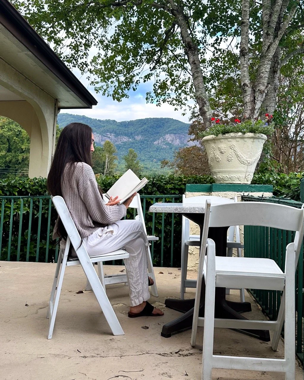 A woman with long dark hair is sitting on a white chair on a porch, reading a book. She is wearing light-colored pants, a gray sweater, and dark sandals. There is a small white table and another white chair on the porch. A large tree with a decorativ