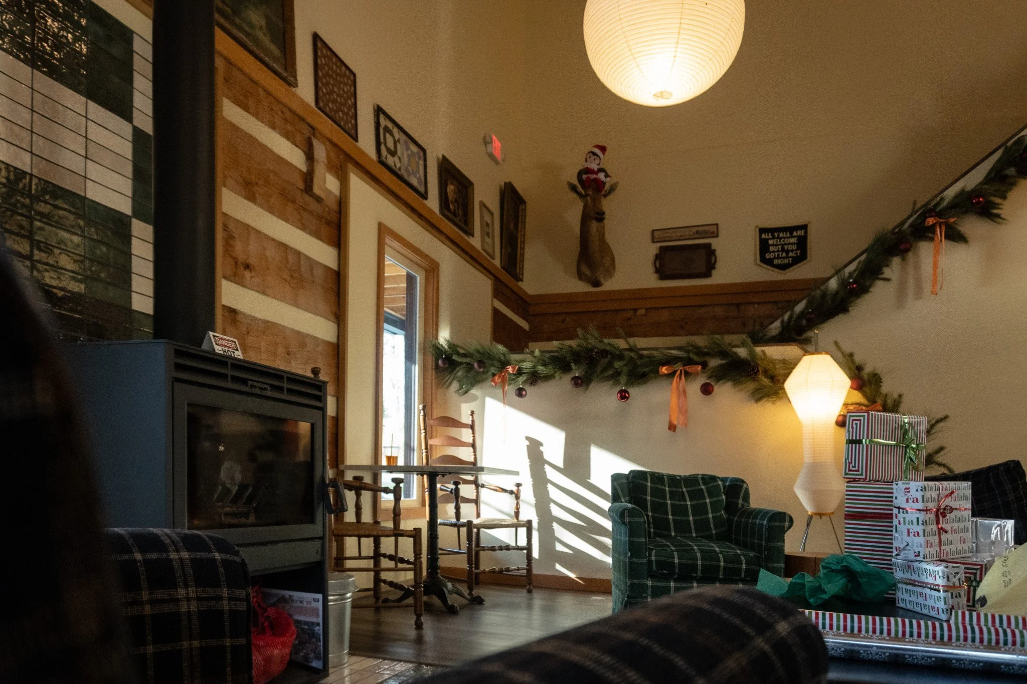 Cozy living room decorated for Christmas with a garland and ornaments, a lamp, and a gift box. There is artwork on the wall, a mounted reindeer head with a Santa hat, and furniture including chairs and a table. Sunlight is streaming in through a wind