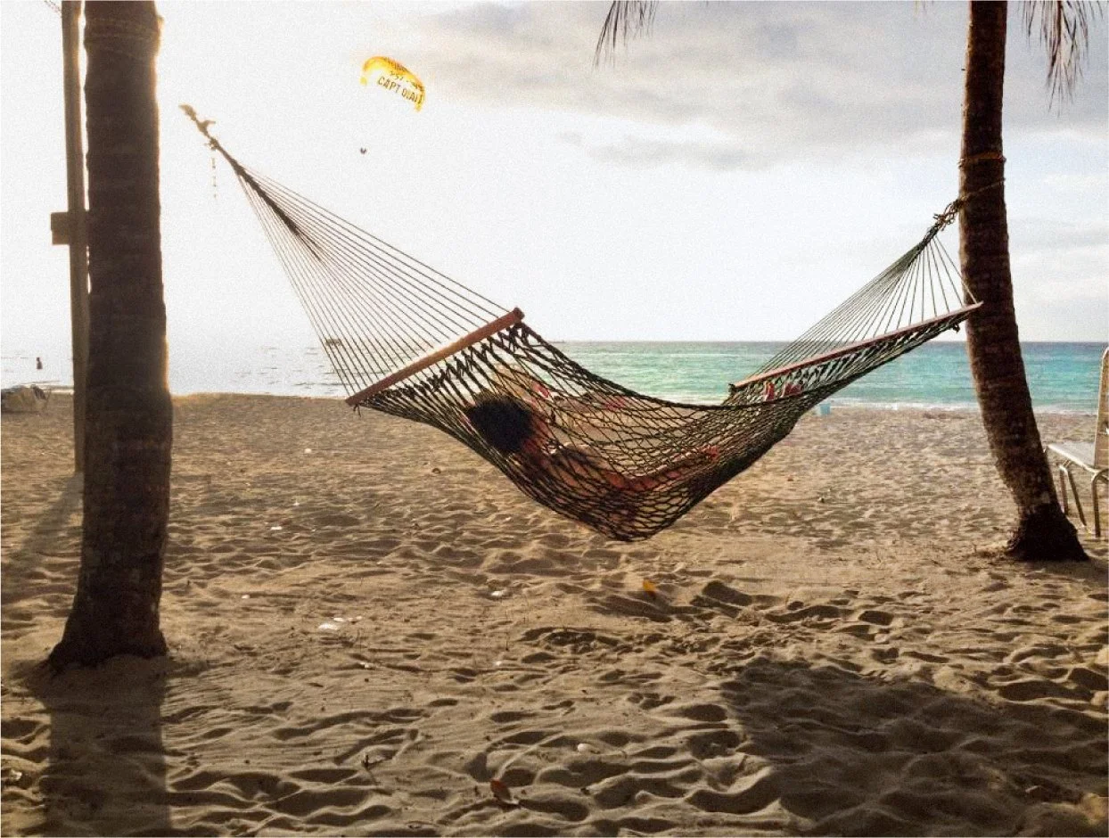 Person lying in a hammock between two palm trees on a sandy beach, with the ocean and cloudy sky in the background.
