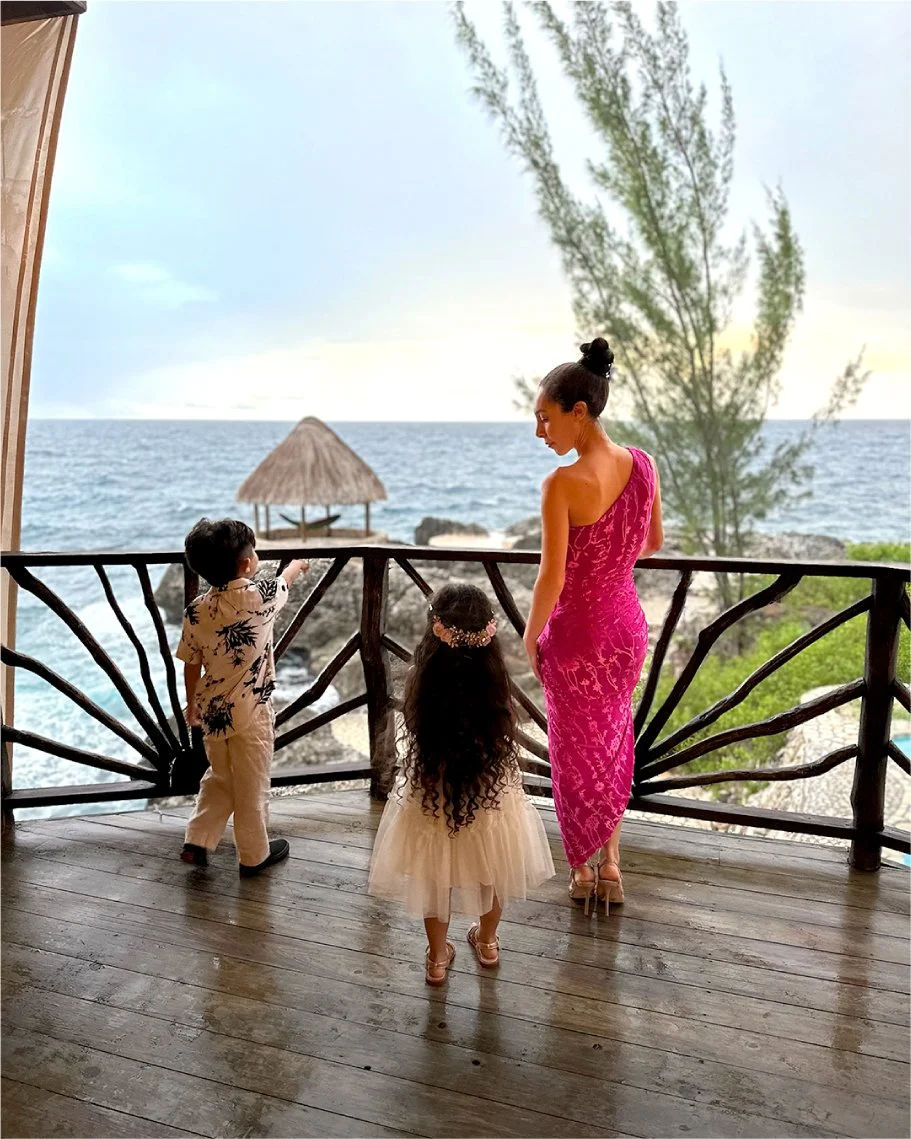 A woman and two young children standing on a wooden balcony overlooking the ocean and a thatched hut on rocks. The woman is wearing a form-fitting pink dress, and the children are dressed in light-colored clothing, one with a floral headband. They ap