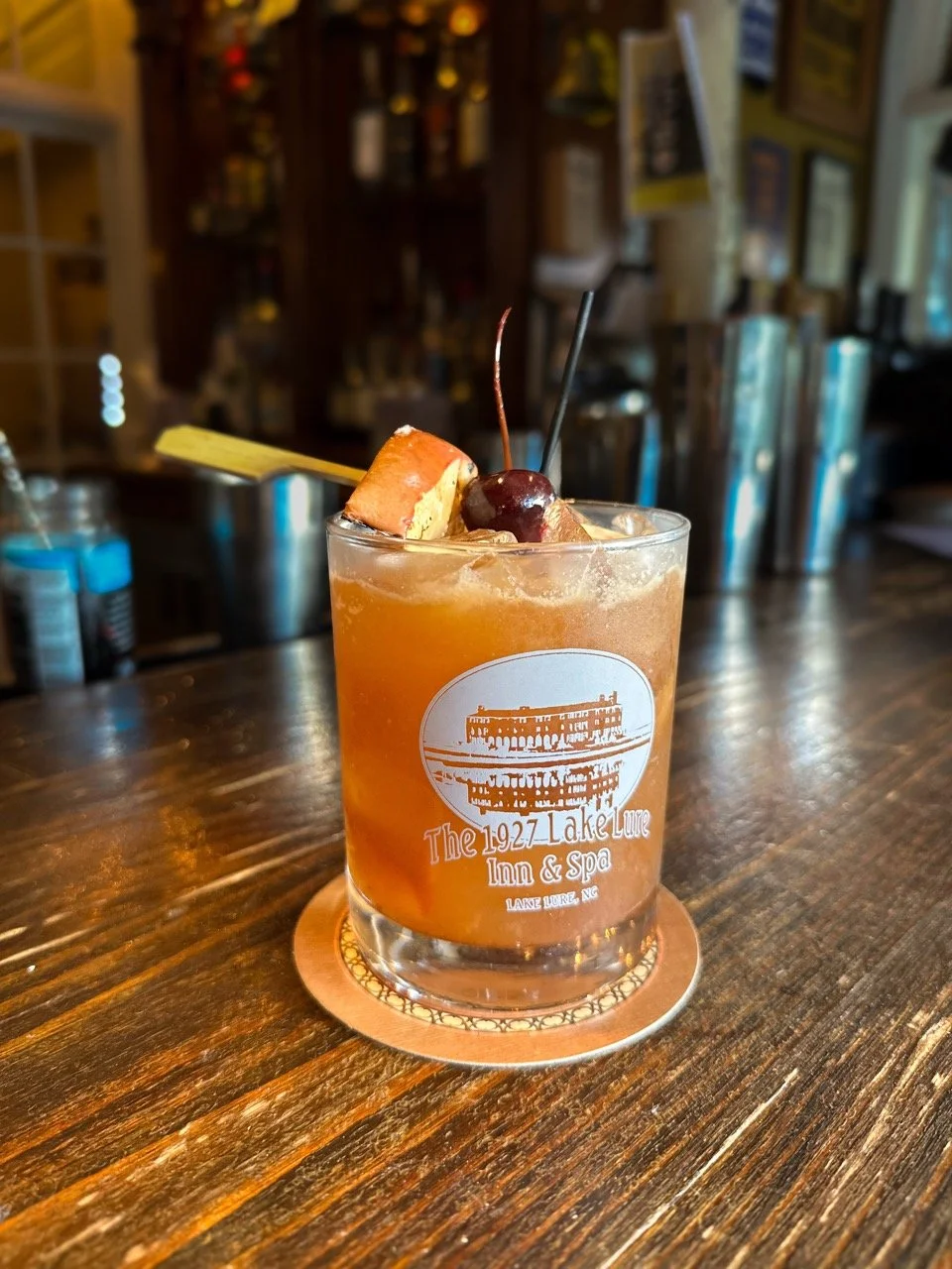An orange cocktail in a glass with ice, garnished with cherries, an orange slice, and a yellow paper umbrella, placed on a wooden bar counter.